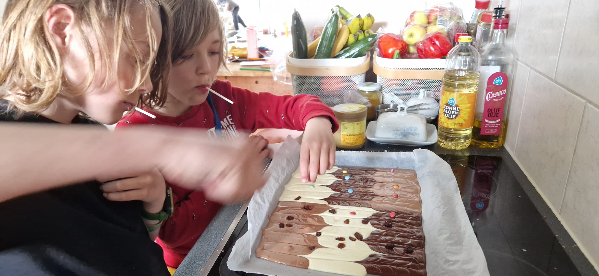 2 jongens staan met een lollie in hun mond in de keuken. Ze maken zelf een lekkernij van chocolade