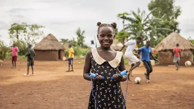 De negenjarige Amina staat in een open vlakte in haar vluchtelingenkamp in Oeganda. Ze houdt een springtouw vast, terwijl achter haar kinderen voetballen